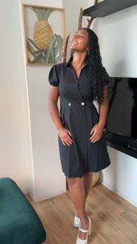 Juodas suknelė su ilgomis pūstomis rankovėmis, liemeniu surištas diržas ir auksiniai mygtukai. Suderinta su baltomis platforminėmis avalynėmis, nustatyta šviesioje patalpoje.