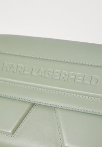 Grønn skinnlommebok med teksturert søm, preget "KARL LAGERFELD"-logo og geometriske mønstre. Glatt overflate, rektangulær form.