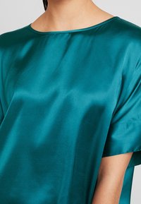 Blouse en satin bleu-vert avec une coupe décontractée, un col large et des manches courtes. Le tissu a une texture lisse et légèrement réfléchissante.