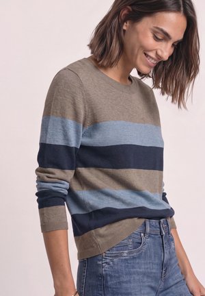 Femme souriante, portant un pull à rayures beige, bleu clair et bleu foncé à manches longues et un jean bleu, debout devant un fond clair.