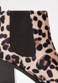 Botín de tobillo de estampado de leopardo beige con parte superior de tejido texturizado, un panel elástico negro y un tacón grueso negro.