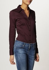 Camisa burdeos de manga larga con cuello puntiagudo, frente abotonado y textura suave, combinada con jeans ajustados de mezclilla azul.