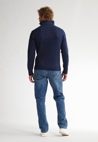 Marineblauwe gebreide trui met een hoge kraag, met gesprenkelde texturen en ribbels aan de zomen, gecombineerd met lichtblauwe jeans en witte sneakers.