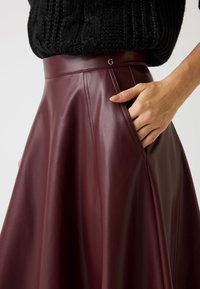 Gonna in pelle bordeaux con vita alta, dotata di tasche laterali e dettagli cuciti. Texture liscia e lucentezza sottile.