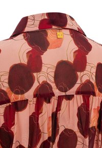 Blouse fluide en tissu léger ; présente un fond rose avec de grands motifs floraux rouge foncé et une bordure dorée sur le col.