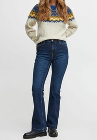 Siniset korkea vyötäröiset leveälahkeiset farkut, joissa on sileä denimituntu, yhdistettynä kermaiseen neuleeseen, jossa on värikkäitä geometrisia kuvioita. Mustat nilkkurit.