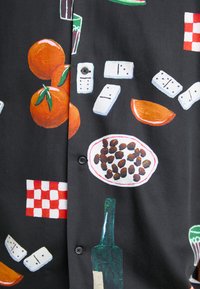 Camisa de manga corta negra con gráficos coloridos de comida y bebida, incluyendo naranjas, cuencos, dominós y una botella verde, con un cierre de botones al frente.