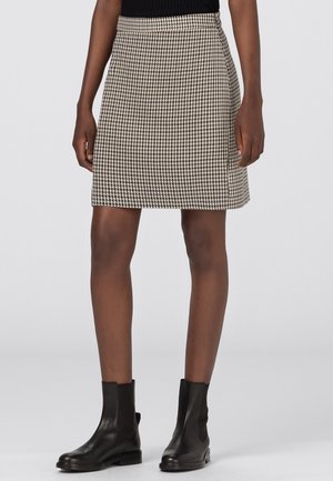 Nainen mustassa topissa, beigen ja mustan houndstooth-kuvioisessa hameessa sekä mustissa nahkaisissa nilkkureissa, seisomassa vaaleaa yksiväristä taustaa vasten.