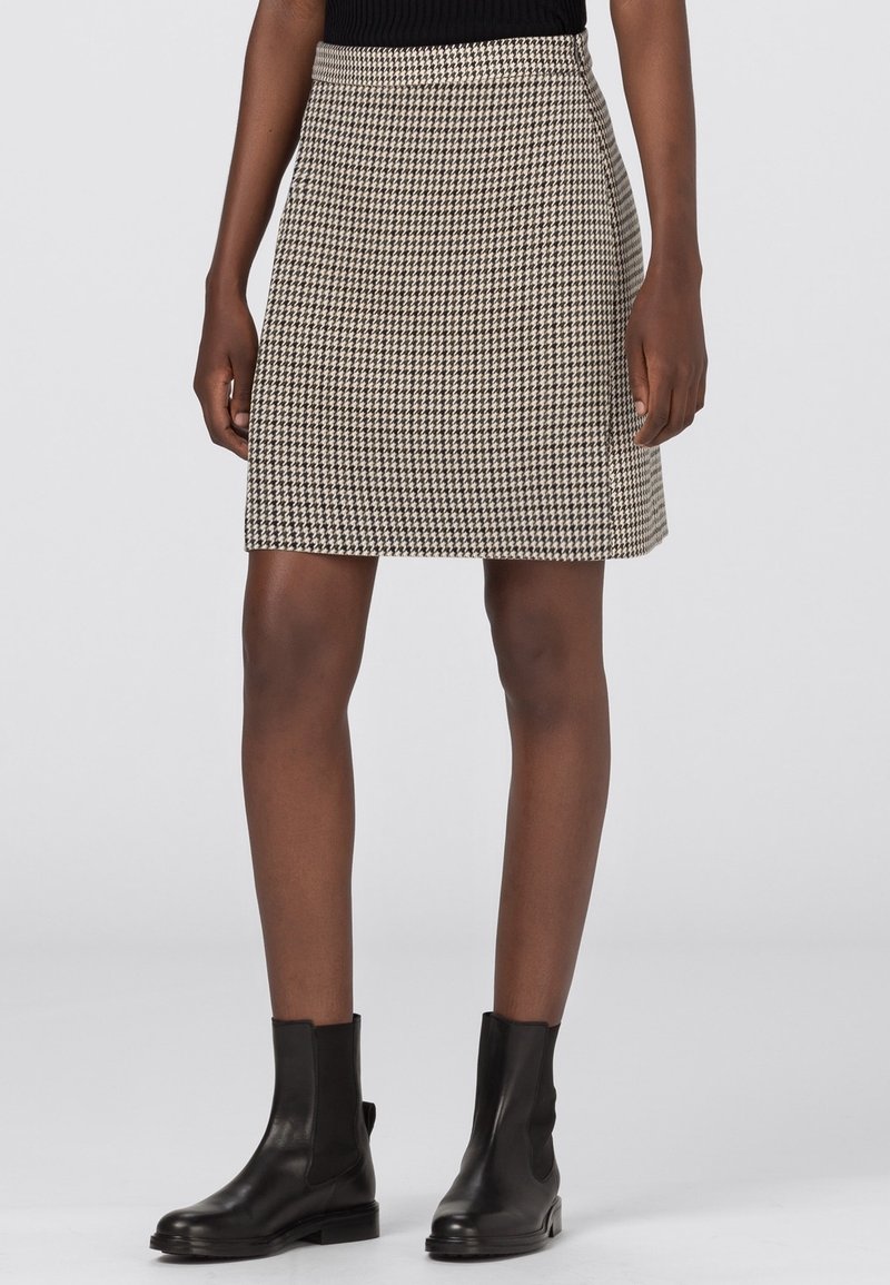 Femme portant un haut noir, une jupe pied-de-poule beige et noire, et des bottines en cuir noir, debout devant un fond clair uni.