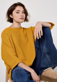 Jeune femme aux cheveux courts châtain clair portant un pull texturé de couleur jaune moutarde, un jean large bleu et des bottes en suede beige, assise avec un genou relevé.