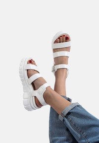 Sandali bianchi chunky con cinture larghe e una fibbia, caratterizzati da una suola in gomma testurizzata, poggiati su uno sfondo neutro.