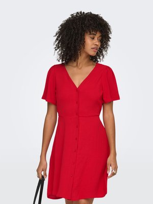 Donna con capelli ricci che indossa un vestito rosso con bottoni sul davanti e maniche corte, tiene le cinghie nere della borsa, guardando verso sinistra.