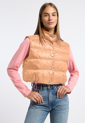 Perzikkleurige, korte puffer vest met een hoge kraag, drukknoopjes en een verstelbare trekkoord aan de onderkant. Ggedragen over een roze langemouwenshirt.