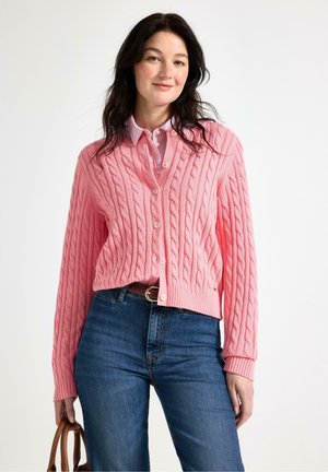Donna con capelli scuri che indossa un cardigan rosa a trecce sopra una camicia a righe, jeans blu, e tiene una borsa marrone su sfondo bianco.
