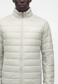 Personne portant une veste matelassée gris clair avec une fermeture éclair à l'avant et le logo EA7 Emporio Armani sur la partie supérieure gauche de la poitrine.