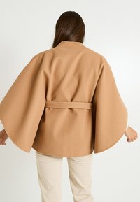 Capa de mezcla de lana color tostado con mangas anchas y cinturón de lazo, que presenta una textura suave. La vista trasera muestra la costura del hombro y un ligero drapeado.