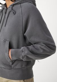 Personne portant un sweat à capuche gris foncé zippé, mains dans la poche avant, manches visibles et poignets côtelés, sur un fond clair uni.