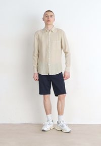 Camicia in lino beige chiaro con taschino, abbottonata, abbinata a pantaloni corti blu navy. Il modello indossa calze bianche e sneakers grigie. Design semplice e rilassato.