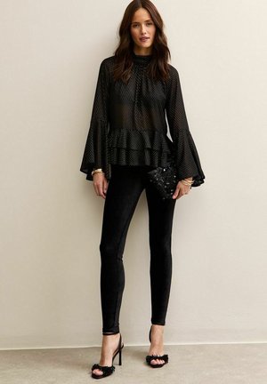 Mujer vestida con una blusa negra transparente de manga larga con puños acampanados y lunares, leggings negros, sandalias de tacón alto, sosteniendo un bolso de lentejuelas negras.