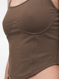 Haut marron ajusté sans manches avec des détails de coutures courbes porté sur le haut du torse sur un fond clair uni.