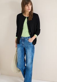 Femme portant un cardigan noir boutonné par-dessus un haut vert clair et un jean bleu, tenant un sac beige, debout contre un mur uni.