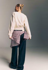 Žena s blond drdolem, oblečená v krémovom svetri, tmavých širokých nohaviciach a prúžkovanej košeli, ktorú má previazanú okolo pásu, stojí na jednolatom pozadí.