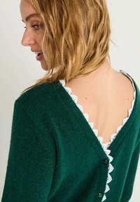 Pull en tricot vert avec un design en V à l'arrière, orné d'une bordure en dentelle blanche et de boutons foncés au dos. Texture douce et coupe décontractée.