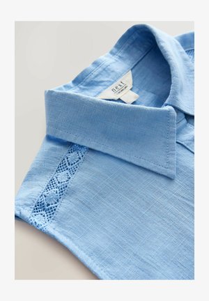 Svijetloplava majica s ovratnikom, koja ima teksturiranu tkaninu i cvjetne čipkaste detalje duž šava na ramenu.