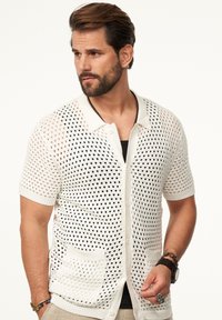 Weißer Strickcardigan mit großem, offenem Netzmuster, kurzen Ärmeln, einem Kragen und zwei Vordertaschen. Der Stoff hat eine leichte Textur.