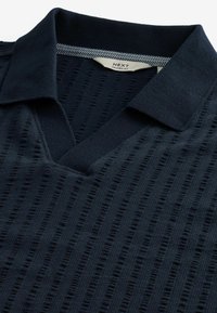 Granatowa, dzianinowa koszula polo z dekoltem w serek, teksturowanym wzorem i kontrastowym wewnętrznym kołnierzykiem. Metka mówi "NEXT Luźny Krój".