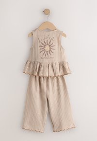 Tenue beige rayée pour tout-petit avec un haut sans manches avec un ourlet à volants et un motif soleil, associée à un pantalon large assorti sur un cintre en bois.