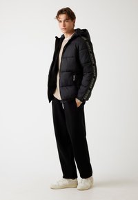Chaqueta de plumas negra con capucha y cremallera, con un detalle de logo en las mangas. Lleva puesta un suéter beige, pantalones de chándal negros y zapatillas blancas.