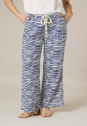 Femme portant un pantalon large à rayures zébrées bleues et blanches avec une ceinture en corde, un haut blanc et des sandales à talons blocs ouverts dorées.