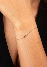 Guldband med en delikat kedja, med två avlånga länkar och intrikata detaljer på ytan. Buren på ett ljust skinnat handled.