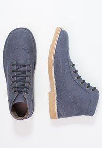 Bottines en tissu bleu avec des lacets noirs, une semelle en caoutchouc beige et des coutures blanches contrastantes. Bout rond, design structuré et six œillets.