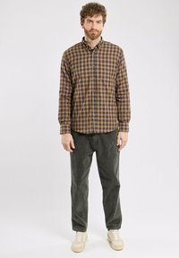 Un homme aux cheveux bouclés et à la barbe se tient portant une chemise à carreaux marron et marine, un pantalon gris foncé et des baskets beige clair sur fond blanc.