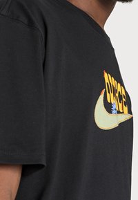 Svart bomull t-shirt med en färgglad broderad logotyp. Designen inkluderar gula och blå detaljer med en grön swoosh.