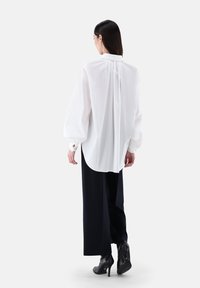 Weiße Bluse mit langen Ärmeln und gerafftem Rücken, kombiniert mit schwarzen, weiten Hosen und hohen schwarzen Stiefeln. Glatte, entspannte Passform.
