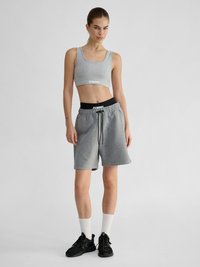 Donna in piedi che indossa un crop top grigio chiaro, pantaloncini grigi con coulisse, calzini bianchi e scarpe da ginnastica nere su sfondo chiaro uniforme.