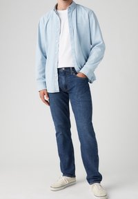 Hombre con camisa de mezclilla azul claro sobre una camiseta blanca, jeans oscuros, zapatillas blancas, con una mano en el bolsillo frente a un fondo liso.