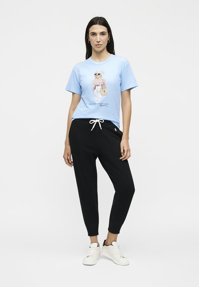 Γυναίκα με ανοιχτό μπλε μπλουζάκι με γραφικό αρκούδας, μαύρο παντελόνι με κορδόνι και λευκά αθλητικά παπούτσια, στέκεται μπροστά σε απλό φόντο.