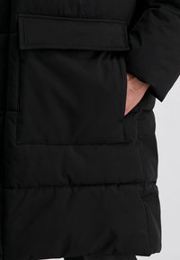Schwarze Pufferjacke mit sichtbarer aufgesetzter Tasche, glatter Textur und gestepptem Detail am Saum, mit einer lockeren Passform.