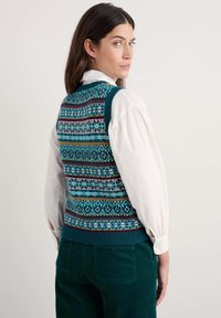 Il gilet lavorato a maglia presenta una base turchese con motivi geometrici multicolori. Indossato sopra una camicia bianca con colletto, abbinato a pantaloni in corduroy verde aderenti.