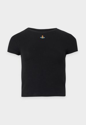 Kvinde med langt sort hår iført en sort tætsiddende t-shirt og løstsiddende lyse beige jeans, der står med den ene hånd i lommen på en ensfarvet hvid baggrund.