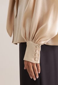 Blusa beige in satin a maniche lunghe con maniche voluminose, caratterizzata da un polsino ornato da quattro bottoni rotondi. Tessuto liscio e tessuto fluente.