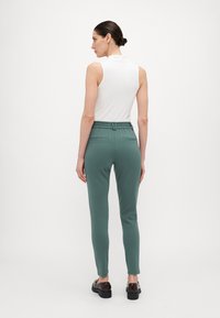 Groene, aansluitende broek met een textuur-elastische tailleband en twee achterzakken, gecombineerd met een witte mouwloze top, gezien van achteren.