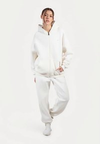 Sudadera blanca con cremallera, capucha grande y bolsillos frontales, combinada con pantalones deportivos ajustados a juego y zapatillas blancas. Tela suave y lisa.