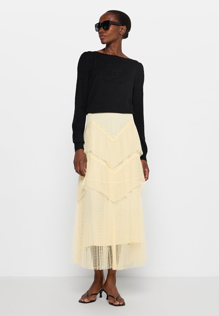 Schwarzer Pullover mit langen Ärmeln und strukturierten Details, kombiniert mit einem tierierten, hellgelben Midirock mit horizontalen Streifen und einem fließenden Saum.