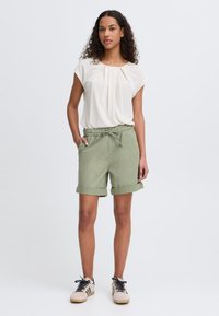 Lys beige kortermet bluse kombinert med olivengrønne shorts med snøring. Shortsene har oppbrettede kanter og sidelommer.