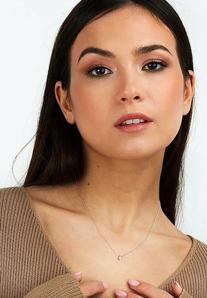 Silberne Halskette mit einem kleinen runden Anhänger, getragen auf einem nackten Schlüsselbein, vor einem strukturierten braunen Pullover mit V-Ausschnitt.